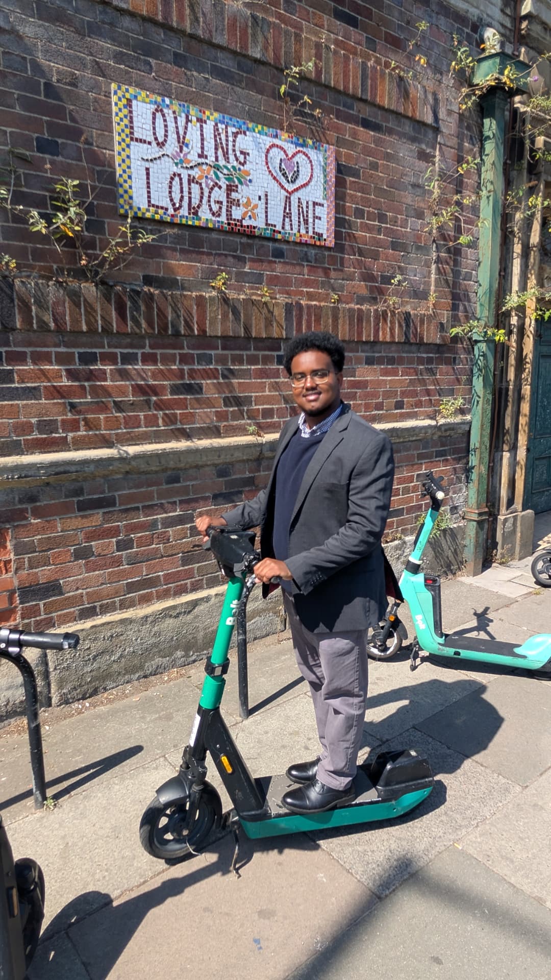 Abdi Ahmed on an electric scooter in front of the Loving Lodge Lane mosaic in Princes Park, L8