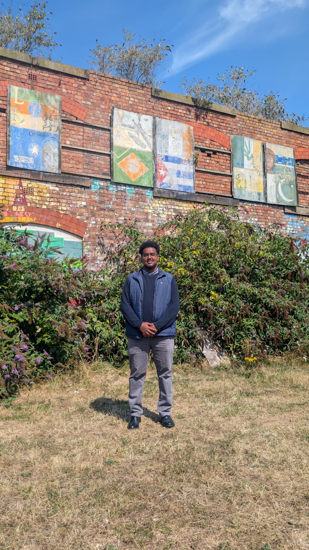 Abdi Ahmed standing in front of a colourful community mural on a brick wall in Princes Park, L8