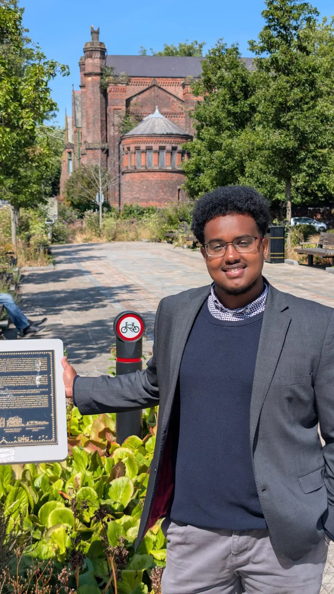 Abdi Ahmed in Princes Park, L8, standing beside a heritage plaque with a historic red-brick building behind him